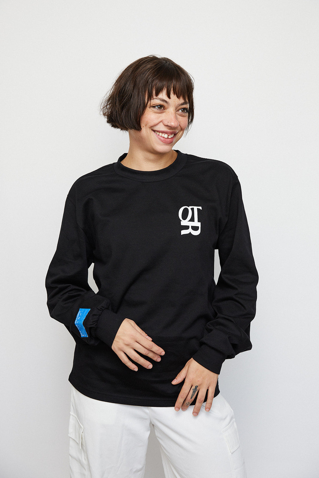 Female model Danielle wearing the "CHANGE YOUR MIND" T-Shirt in unisex size Small and the color black. Danielle is facing the camera, smiling. There is an OTR logo on the chest and a blue woven tag on the right arm, just near the cuff.